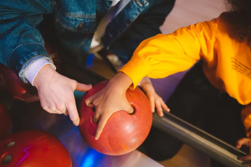 La main d'un adulte et celle d'un enfant posées sur une boule de bowling rouge, au retour de boules