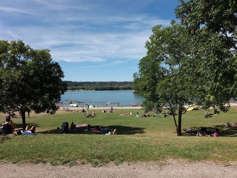 Des baigneurs allongés sur une pelouse en pente douce qui descend vers le lac des Eaux Bleues, sous des arbres, un jour d'été